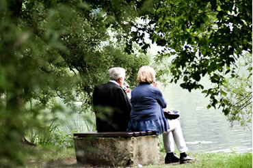 Older man and woman sitting beside a pond under trees